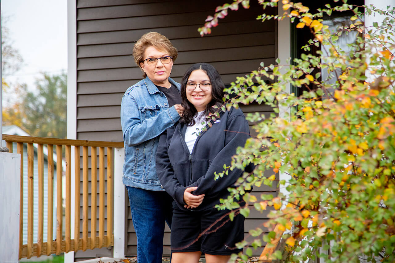 A woman stands with her arm resting on her teenage daughter's shoulder, as they smile on the front porch of their home.